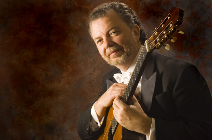 Manuel Barrueco in white tie, holding his guitar, lit warmly against a mottled dark red ground