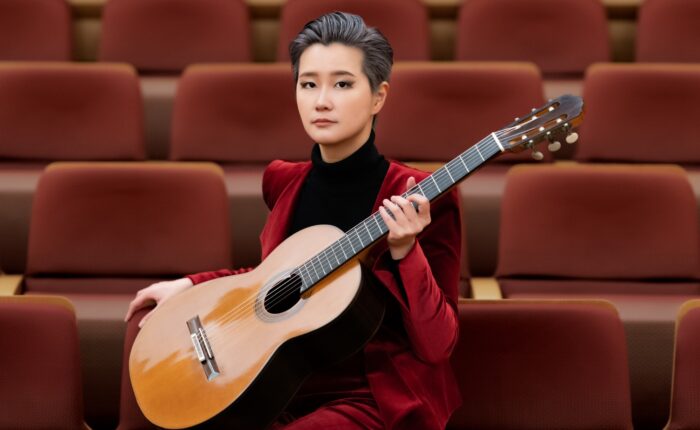 Meng Su seated in a hall of red velvet seats, guitar across her lap, wearing a deep red suit
