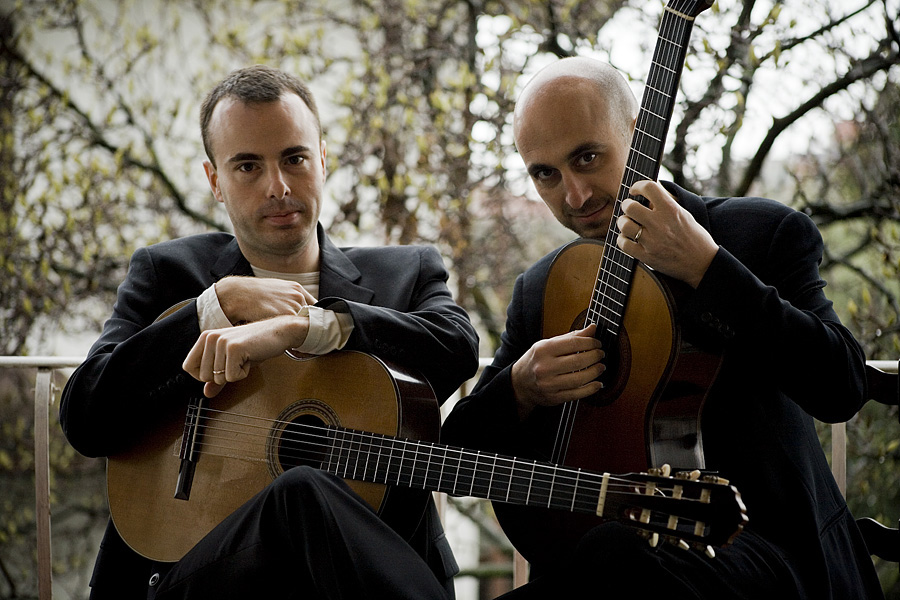 SoloDuo, two guitarists seated outdoors with their instruments, in dark suits against bare spring branches