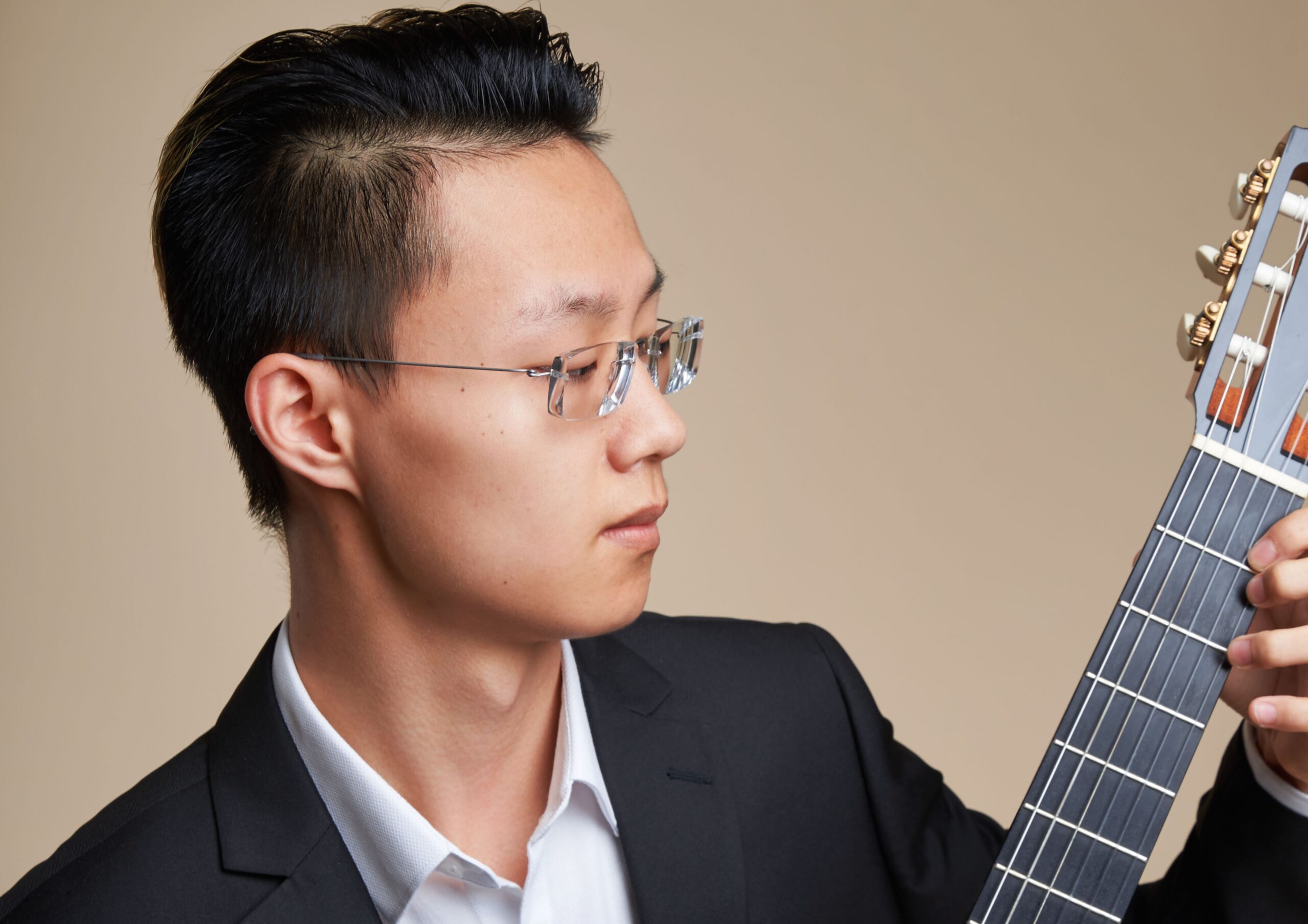 A young guitarist in a dark suit studying the fretboard of his instrument in profile