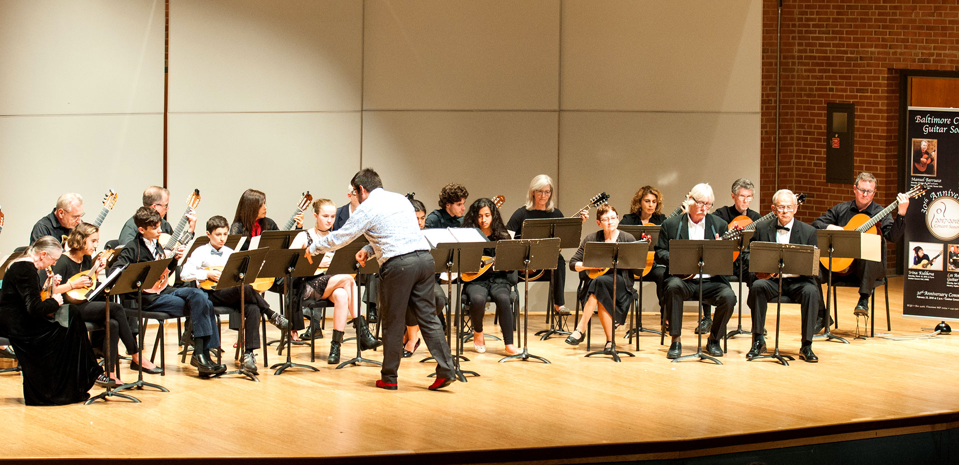 The BCGS Guitar Orchestra performing on stage, conductor mid-gesture before two rows of seated players